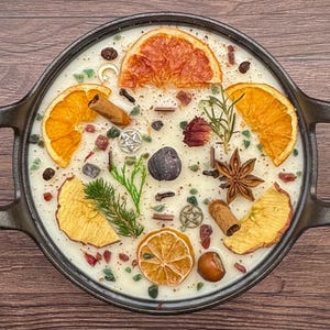 May include: A decorative candle in a black cast iron pan. The candle is white and adorned with orange slices, star anise, cinnamon sticks, and other botanicals. The pan has two handles and sits on a wooden surface.