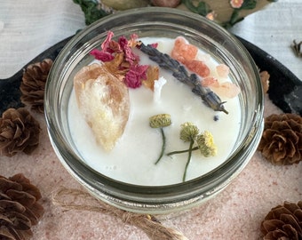 Puede incluir: Una vela blanca en un tarro de vidrio transparente con tapa. La vela está decorada con lavanda seca, sal rosa del Himalaya, flores secas y un cristal de cuarzo transparente. El tarro está sentado en una cama de sal rosa del Himalaya.