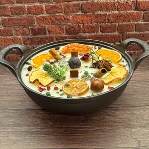 May include: A black cast iron candle with a creamy white wax base, adorned with dried orange slices, cinnamon sticks, and star anise. The candle is decorated with small green and red stones, and a sprig of greenery. The candle has two handles.