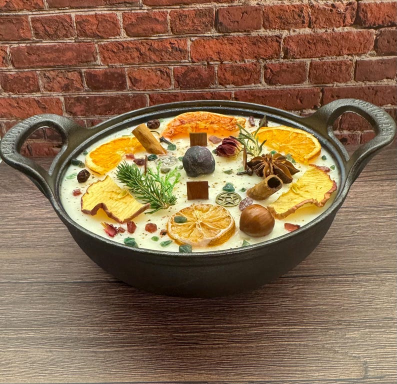 May include: A black cast iron candle with a creamy white wax base, adorned with dried orange slices, cinnamon sticks, and star anise. The candle is decorated with small green and red stones, and a sprig of greenery. The candle has two handles.