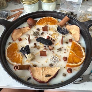 Puede incluir: Una vela grande y redonda en una sart&eacute;n de hierro fundido negro, decorada con rodajas de naranja secas, rodajas de manzana, palitos de canela y otros elementos bot&aacute;nicos. La vela tiene una base de cera blanca.