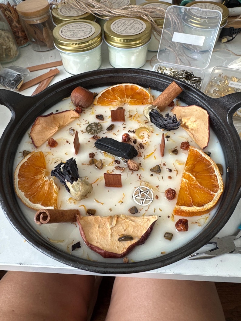 Puede incluir: Una vela grande y redonda en una sart&eacute;n de hierro fundido negro, decorada con rodajas de naranja secas, rodajas de manzana, palitos de canela y otros elementos bot&aacute;nicos. La vela tiene una base de cera blanca.