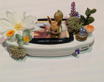 Puede incluir: Una consola portátil Game Boy Advance blanca decorada con flores artificiales, suculentas verdes, pequeños hongos coloridos y una figura marrón de Eevee colocada sobre la pantalla, que muestra una carta coleccionable de Eevee.