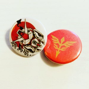Pode incluir: Dois bot&otilde;es redondos vermelhos e brancos com designs inspirados em anime. Um bot&atilde;o apresenta uma cabe&ccedil;a de rob&ocirc; vermelha e branca com texto japon&ecirc;s. O outro bot&atilde;o apresenta um s&iacute;mbolo alado dourado em um fundo vermelho.