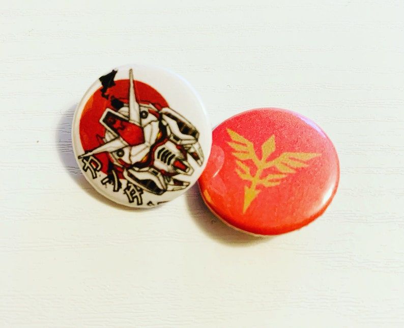 Pode incluir: Dois bot&otilde;es redondos vermelhos e brancos com designs inspirados em anime. Um bot&atilde;o apresenta uma cabe&ccedil;a de rob&ocirc; vermelha e branca com texto japon&ecirc;s. O outro bot&atilde;o apresenta um s&iacute;mbolo alado dourado em um fundo vermelho.