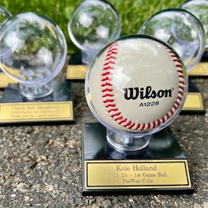 May include: A baseball encased in a clear display dome, mounted on a black base with a gold nameplate. The baseball is white with red stitching and the Wilson logo. The nameplate reads "Kole Holland 5.11.24 - 1st Game Ball PeeWee Cubs."
