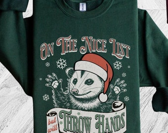 May include: Two Christmas themed sweatshirts, one maroon and one forest green, feature a possum wearing a Santa hat and the text "On the Nice List but will Throw Hands".