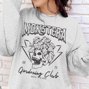 May include: Ash-colored sweatshirt with a black graphic featuring the word "MONSTERA" above a skull and plant design. Below the design is "Gardening Club" and "Rooted in Chaos". The sweatshirt is a crew neck style.