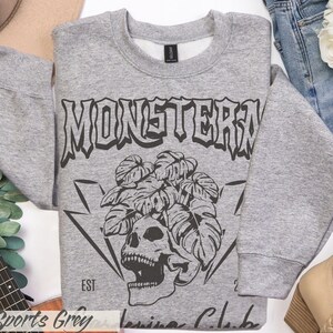 May include: Sports grey sweatshirt with a black graphic of a skull, leaves, and the word "MONSTERA". The sweatshirt has a crew neck and long sleeves. The text "EST 2021" is also visible.