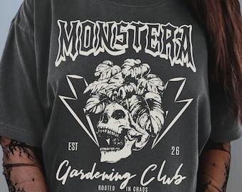 Peut inclure: Un t-shirt gris anthracite à manches courtes avec un motif blanc représentant un crâne surmonté de feuilles de monstera, encadré par des éclairs. Le texte indique MONSTERA, EST 26, Gardening Club et ROOTED IN CHAOS.
