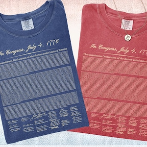 May include: Two Comfort Colors t-shirts, one denim blue and one crimson red, are displayed. Each shirt features the text "In Congress, July 4, 1776" and the Declaration of Independence. The shirts are labeled "Denim" and "Crimson".