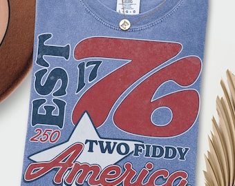 May include: Blue jean colored t-shirt with a distressed graphic. The graphic includes the text "EST 17 76 250 TWO FIDDY America" in red, white, and blue. A gold necklace is draped over the shirt.