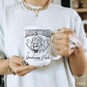 May include: White ceramic mug with the words "MONSTERA Gardening Club" in black, along with a skull and plant graphic. The mug is held by a person wearing a gray t-shirt and gold jewelry. The mug is 15oz.