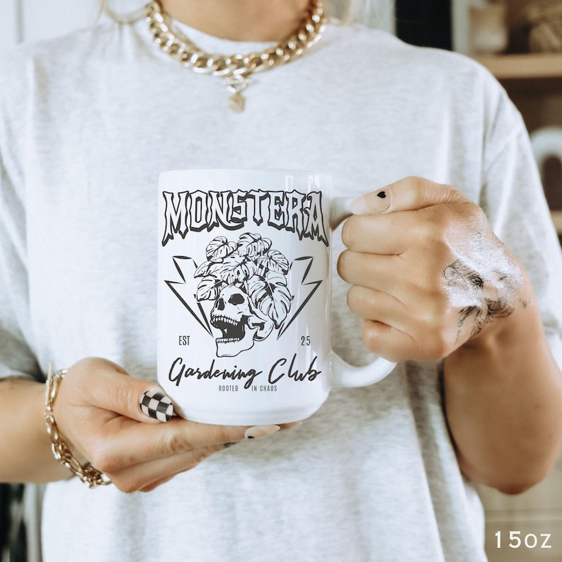 May include: White ceramic mug with the words "MONSTERA Gardening Club" in black, along with a skull and plant graphic. The mug is held by a person wearing a gray t-shirt and gold jewelry. The mug is 15oz.