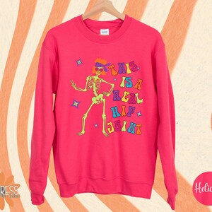 Pu&ograve; includere: Una felpa rosa acceso con una grafica colorata di uno scheletro con occhiali da sole e una fascia. Il testo "THIS IS A REAL HIP JOINT" &egrave; stampato sopra lo scheletro. La felpa &egrave; appesa a una gruccia di legno.