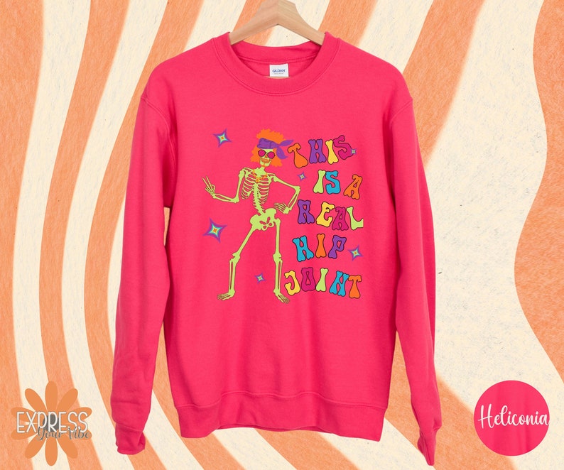 Pu&ograve; includere: Una felpa rosa acceso con una grafica colorata di uno scheletro con occhiali da sole e una fascia. Il testo "THIS IS A REAL HIP JOINT" &egrave; stampato sopra lo scheletro. La felpa &egrave; appesa a una gruccia di legno.