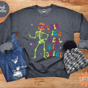 Pu&ograve; includere: Felpa grigio scuro con grafica di scheletro colorata e la scritta "THIS IS A REAL HIP JOINT". Lo scheletro ha capelli arancioni e occhiali da sole. La felpa &egrave; abbinata a jeans e un berretto bianco e nero.