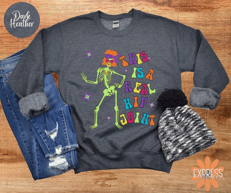 Pu&ograve; includere: Felpa grigio scuro con grafica di scheletro colorata e la scritta "THIS IS A REAL HIP JOINT". Lo scheletro ha capelli arancioni e occhiali da sole. La felpa &egrave; abbinata a jeans e un berretto bianco e nero.