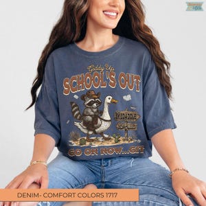 May include: A denim blue t-shirt with a graphic that reads "Giddy Up SCHOOL'S OUT". The graphic features a raccoon in a cowboy hat riding a goose, with a sign that says "Skedaddle 1st Grade". The text "GO ON NOW...GIT" is also included.