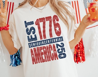 May include: Ivory-colored t-shirt with a patriotic design. The design features the text "EST 1776 SEMIQUINCENTENNIAL AMERICA 250" in red and blue, with stars. The shirt is part of the Comfort Colors collection.