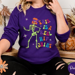 Pu&ograve; includere: Una felpa viola con una grafica di scheletro verde neon. Lo scheletro indossa occhiali da sole e una fascia, con il testo "THIS IS A REAL HIP JOINT" in lettere colorate. La felpa &egrave; di colore unito.