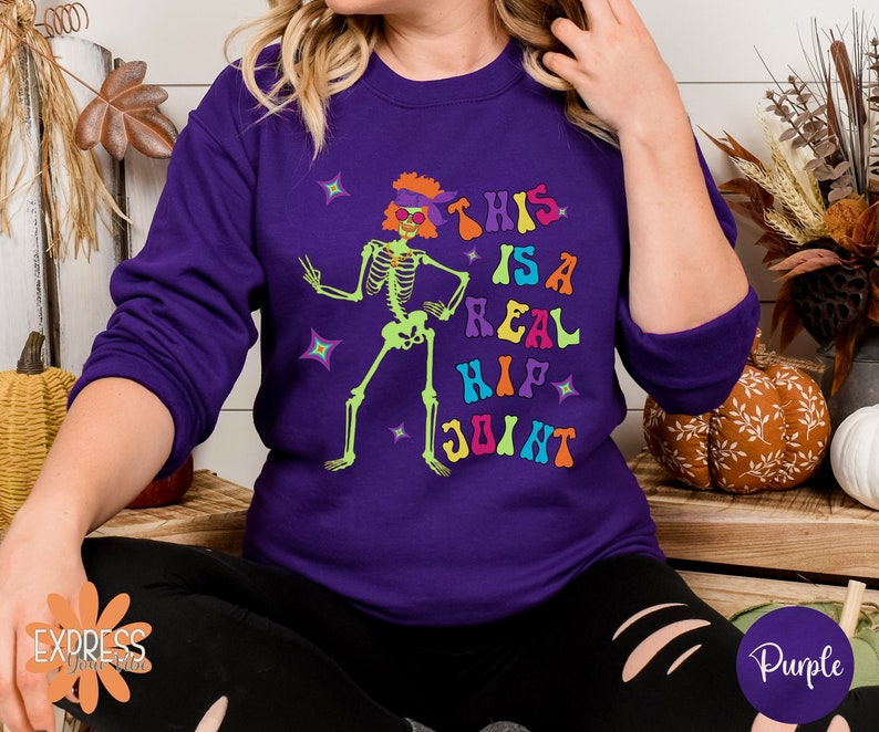 Pu&ograve; includere: Una felpa viola con una grafica di scheletro verde neon. Lo scheletro indossa occhiali da sole e una fascia, con il testo "THIS IS A REAL HIP JOINT" in lettere colorate. La felpa &egrave; di colore unito.