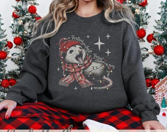 May include: Dark heather grey sweatshirt featuring a possum wearing a red and white plaid scarf with Christmas lights. The text "Festive but Feral!" is printed on the sweatshirt.