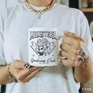 May include: White ceramic mug with the text "MONSTERA Gardening Club" and a skull graphic. The mug has a handle and is labeled "11oz". The design includes a skull with leaves and lightning bolts.