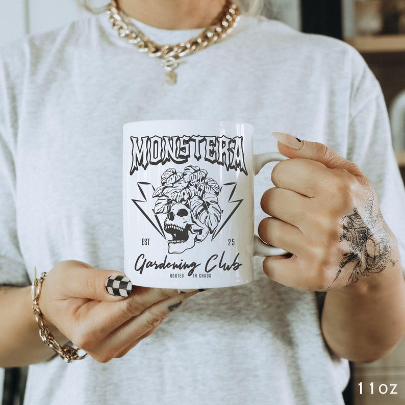 May include: White ceramic mug with the text "MONSTERA Gardening Club" and a skull graphic. The mug has a handle and is labeled "11oz". The design includes a skull with leaves and lightning bolts.