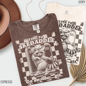 May include: A brown t-shirt with a vintage-style graphic of a raccoon wearing a cowboy hat and boots. The graphic says "Time to Skedadle" and "Go on now...git."