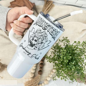 May include: White insulated tumbler with a handle, featuring a black and white graphic of a skull, monstera leaves, and the words "MONSTERA Gardening Club". Includes a silver straw with a white tip. The tumbler is held by a person.
