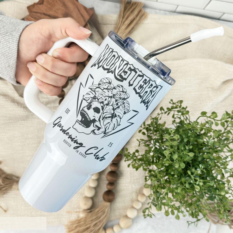 May include: White insulated tumbler with a handle, featuring a black and white graphic of a skull, monstera leaves, and the words "MONSTERA Gardening Club". Includes a silver straw with a white tip. The tumbler is held by a person.