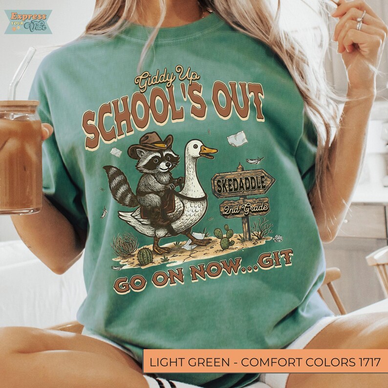 May include: Light green t-shirt with a graphic featuring a raccoon riding a goose, text that reads "School's Out", and the phrase "Go on now...Git". The design includes a sign that says "Skedaddle 2nd Grade".