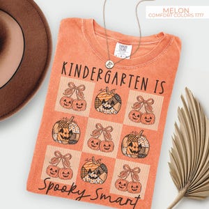 May include: Melon-colored t-shirt with the text "Kindergarten Is Spooky Smart." The shirt features a grid pattern with pumpkin and cherry graphics. A gold necklace with a letter "E" pendant is visible.