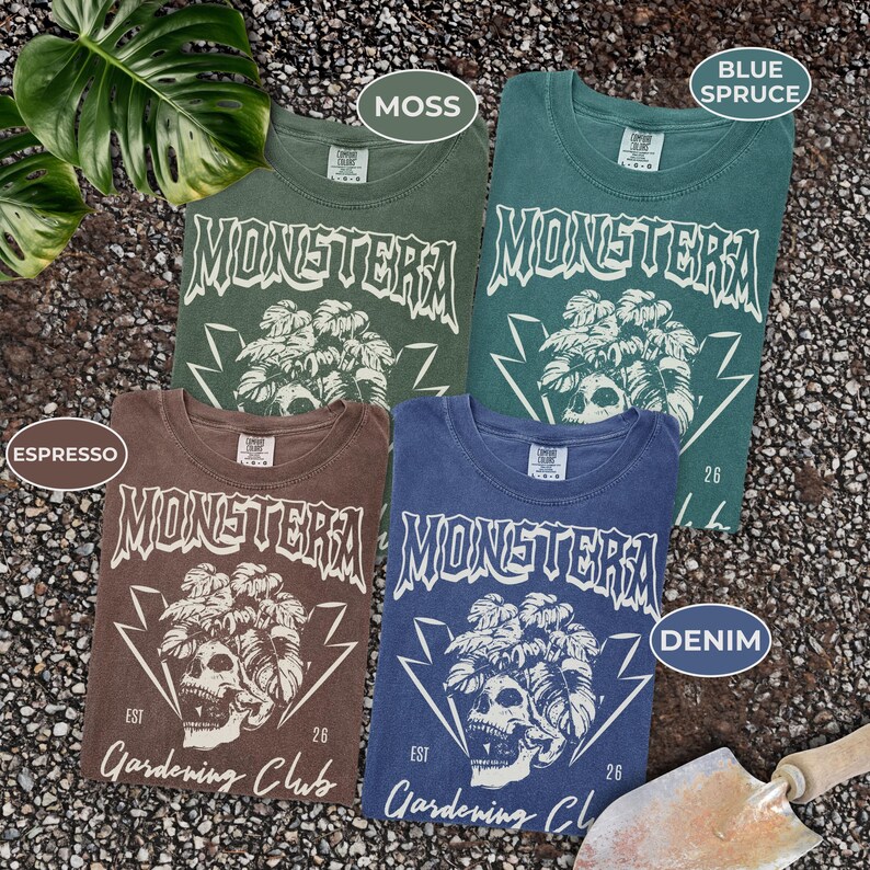 May include: Four t-shirts in moss green, blue spruce, espresso brown, and denim blue. Each shirt features a graphic with the word "MONSTERA", a skull, and the text "Gardening Club". The shirts are folded and displayed on a textured surface.
