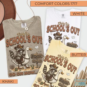 May include: Three Comfort Colors t-shirts in khaki, white, and butter yellow. Each shirt features a graphic with a raccoon wearing a cowboy hat riding a goose, with the text "SCHOOL'S OUT" and "SKEDADDLE".