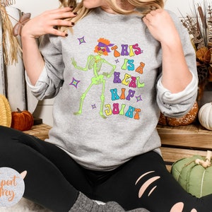 Pu&ograve; includere: Felpa grigio chiaro con una grafica colorata di uno scheletro danzante con occhiali da sole e la frase "THIS IS A REAL HIP JOINT". Il design presenta colori vivaci e stelle, perfetto per Halloween o un look retr&ograve; divertente.