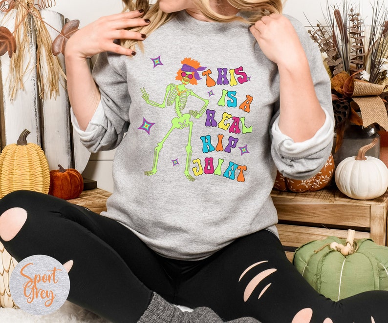 Pu&ograve; includere: Felpa grigio chiaro con una grafica colorata di uno scheletro danzante con occhiali da sole e la frase "THIS IS A REAL HIP JOINT". Il design presenta colori vivaci e stelle, perfetto per Halloween o un look retr&ograve; divertente.