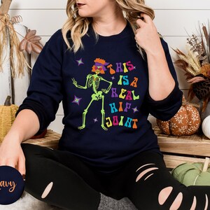 Pu&ograve; includere: Felpa blu navy con la scritta "THIS IS A REAL HIP JOINT" e una grafica di scheletro verde neon. Lo scheletro indossa un cappello e occhiali da sole viola. Design a tema Halloween.