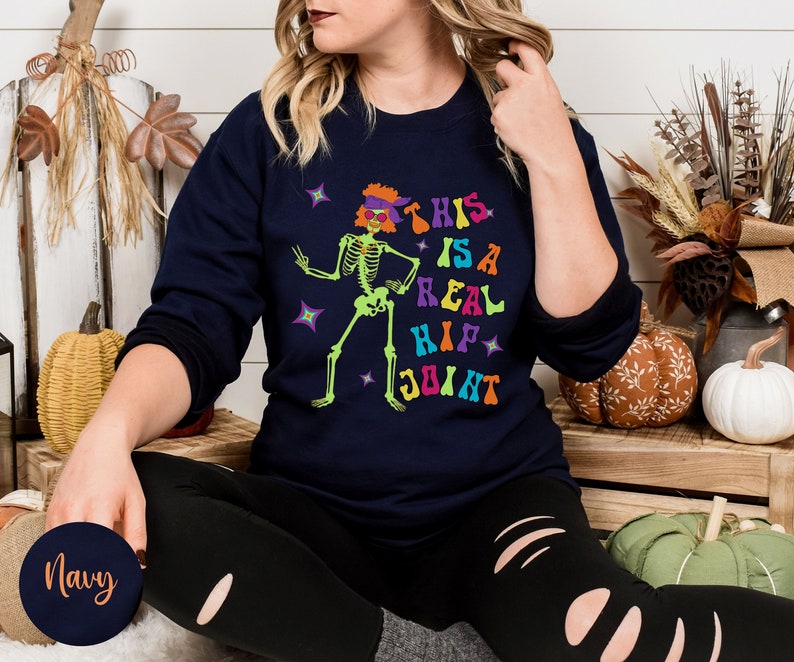 Pu&ograve; includere: Felpa blu navy con la scritta "THIS IS A REAL HIP JOINT" e una grafica di scheletro verde neon. Lo scheletro indossa un cappello e occhiali da sole viola. Design a tema Halloween.