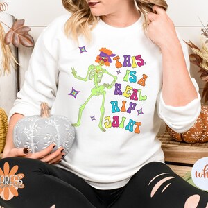 Pu&ograve; includere: Felpa bianca con uno scheletro verde neon con occhiali da sole e una fascia. Il testo "THIS IS A REAL HIP JOINT" &egrave; stampato in lettere colorate. Il design include stelle.