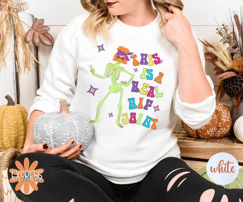 Pu&ograve; includere: Felpa bianca con uno scheletro verde neon con occhiali da sole e una fascia. Il testo "THIS IS A REAL HIP JOINT" &egrave; stampato in lettere colorate. Il design include stelle.