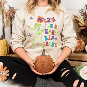 Pu&ograve; includere: Felpa beige con una grafica colorata di uno scheletro danzante e la scritta "THIS IS A REAL HIP JOINT". Lo scheletro &egrave; verde, con una fascia e occhiali da sole viola. Il testo &egrave; in vari colori. Fa parte di una collezione a tema Halloween.
