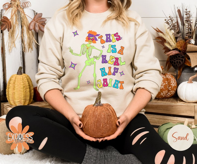 Pu&ograve; includere: Felpa beige con una grafica colorata di uno scheletro danzante e la scritta "THIS IS A REAL HIP JOINT". Lo scheletro &egrave; verde, con una fascia e occhiali da sole viola. Il testo &egrave; in vari colori. Fa parte di una collezione a tema Halloween.