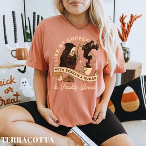 May include: Terracotta-colored t-shirt with a Halloween-themed graphic. The design features a Grim Reaper with a scythe, coffee beans, and the text "I like my coffee with scream & sugar & freshly booed."