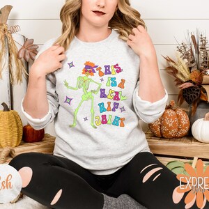 Pu&ograve; includere: Felpa grigio chiaro con una grafica colorata di uno scheletro con occhiali da sole e una fascia. Il testo "THIS IS A REAL HIP JOINT" &egrave; stampato sopra lo scheletro. Il design ha uno stile retr&ograve; e groovy, perfetto per Halloween.