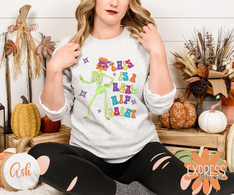 Pu&ograve; includere: Felpa grigio chiaro con una grafica colorata di uno scheletro con occhiali da sole e una fascia. Il testo "THIS IS A REAL HIP JOINT" &egrave; stampato sopra lo scheletro. Il design ha uno stile retr&ograve; e groovy, perfetto per Halloween.
