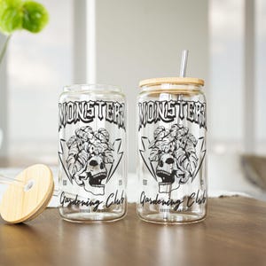 May include: Two clear glass drinking cups with a black and white design. The design includes the text "MONSTER Gardening Club" with a skull and plant graphic. One cup has a bamboo lid and metal straw. The other cup has a clear straw and bamboo lid.