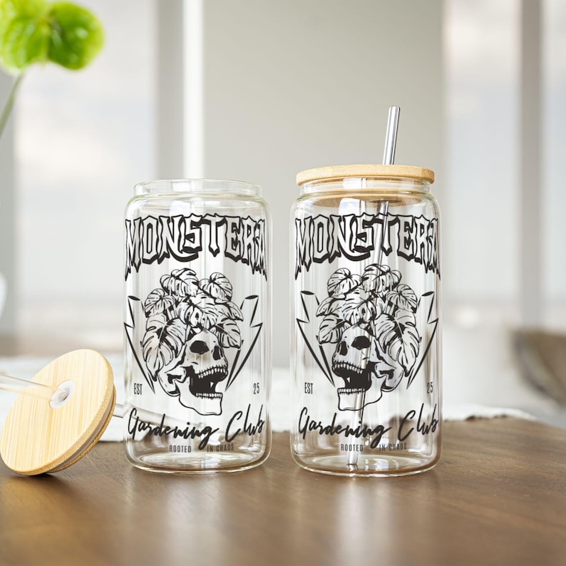 May include: Two clear glass drinking cups with a black and white design. The design includes the text "MONSTER Gardening Club" with a skull and plant graphic. One cup has a bamboo lid and metal straw. The other cup has a clear straw and bamboo lid.