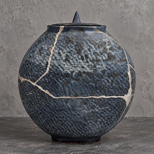 May include: A round, dark blue ceramic vessel with a textured, dimpled surface and a matching lid featuring a pointed finial. The exterior displays prominent, light-colored crack lines across the body, resembling the kintsugi repair technique.